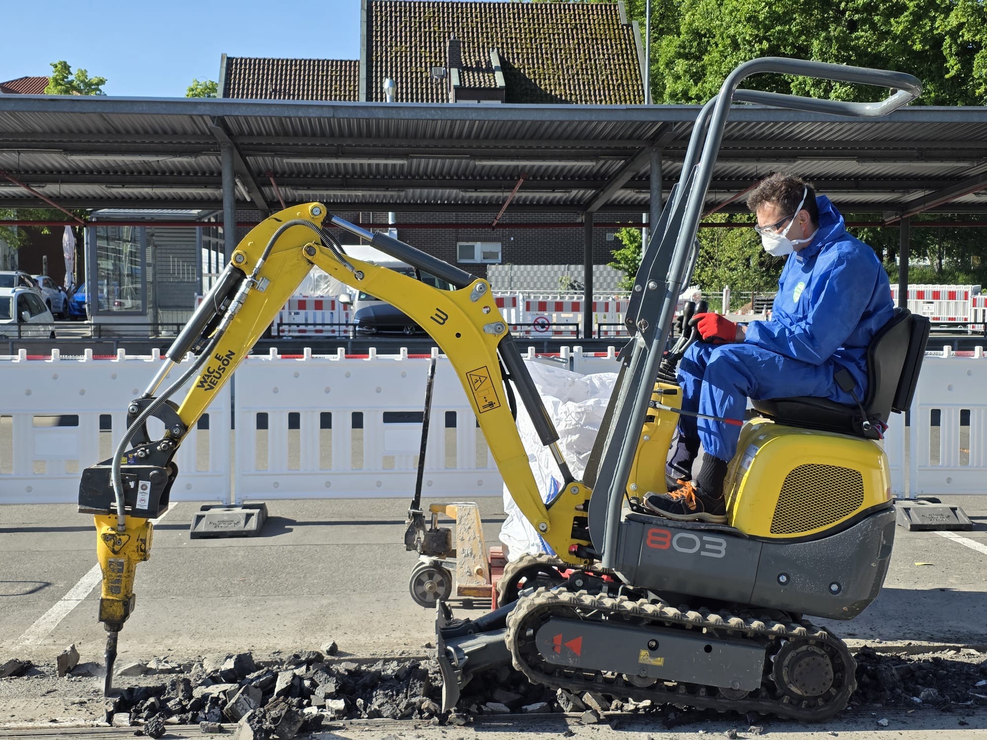Abbruch Asbest Ein gelber Minibagger mit Stemmhammer bei Abbrucharbeiten von Asbest auf einem markierten Außenparkplatz