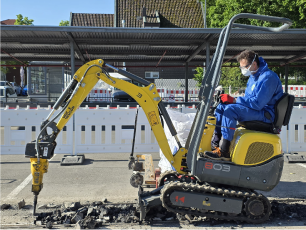 Ein gelber Minibagger mit Stemmhammer bei Abbrucharbeiten von Asbest auf einem markierten Außenparkplatz